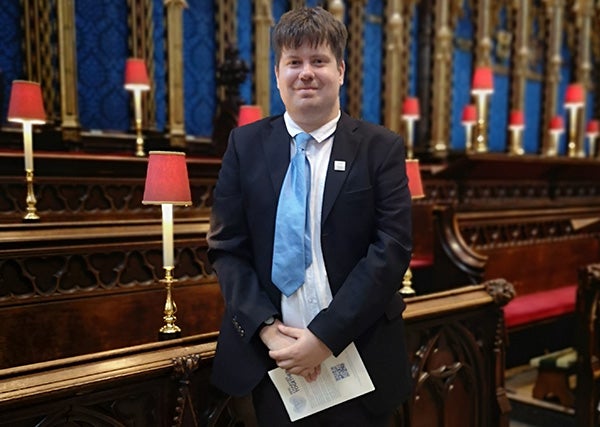 A man with short dark hair, black suit and blue tie standing in Westminster Abbey with rows of seating behind him