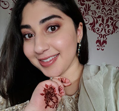 Jameela has dark hair and smiles at the camera. She is holding her hand under her chin, showing henna patterns painted on her palm