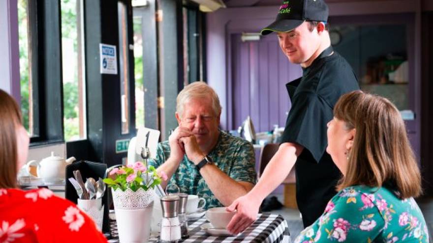A waiter with downs syndrome serving three people at a table in a cafe with coffee