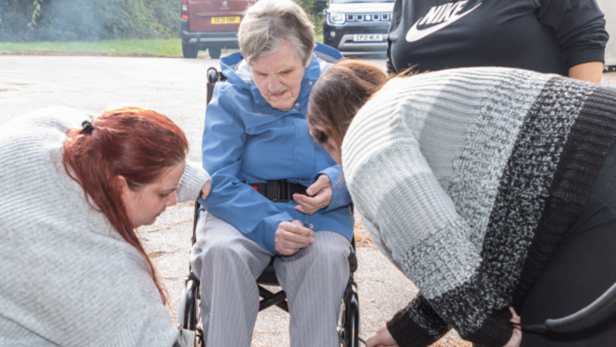 Three support workers are helping a woman in a wheelchair get onto a ramp to get into a mini van