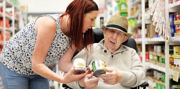 A support worker helping an elderly man in a wheelchair choose groceries in a supermarket