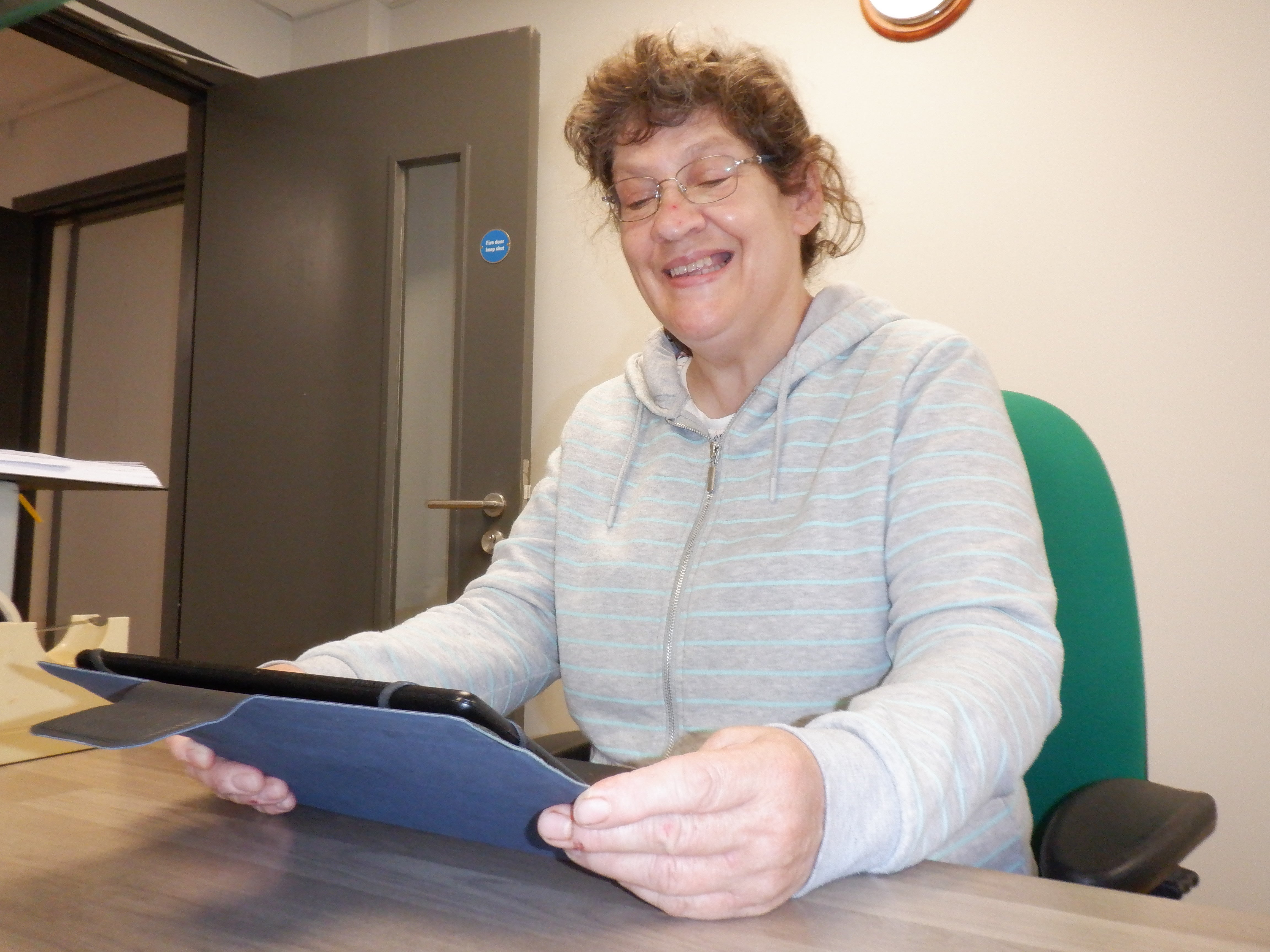 Carol sitting at a desk holding a tablet and smiling to the camera