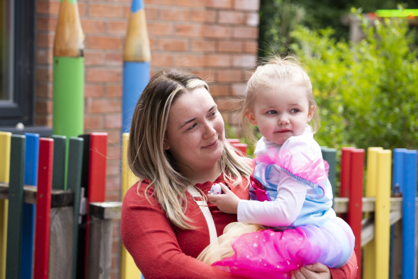 A mother holding a child in her arms and smiling with outside play equipment behind them