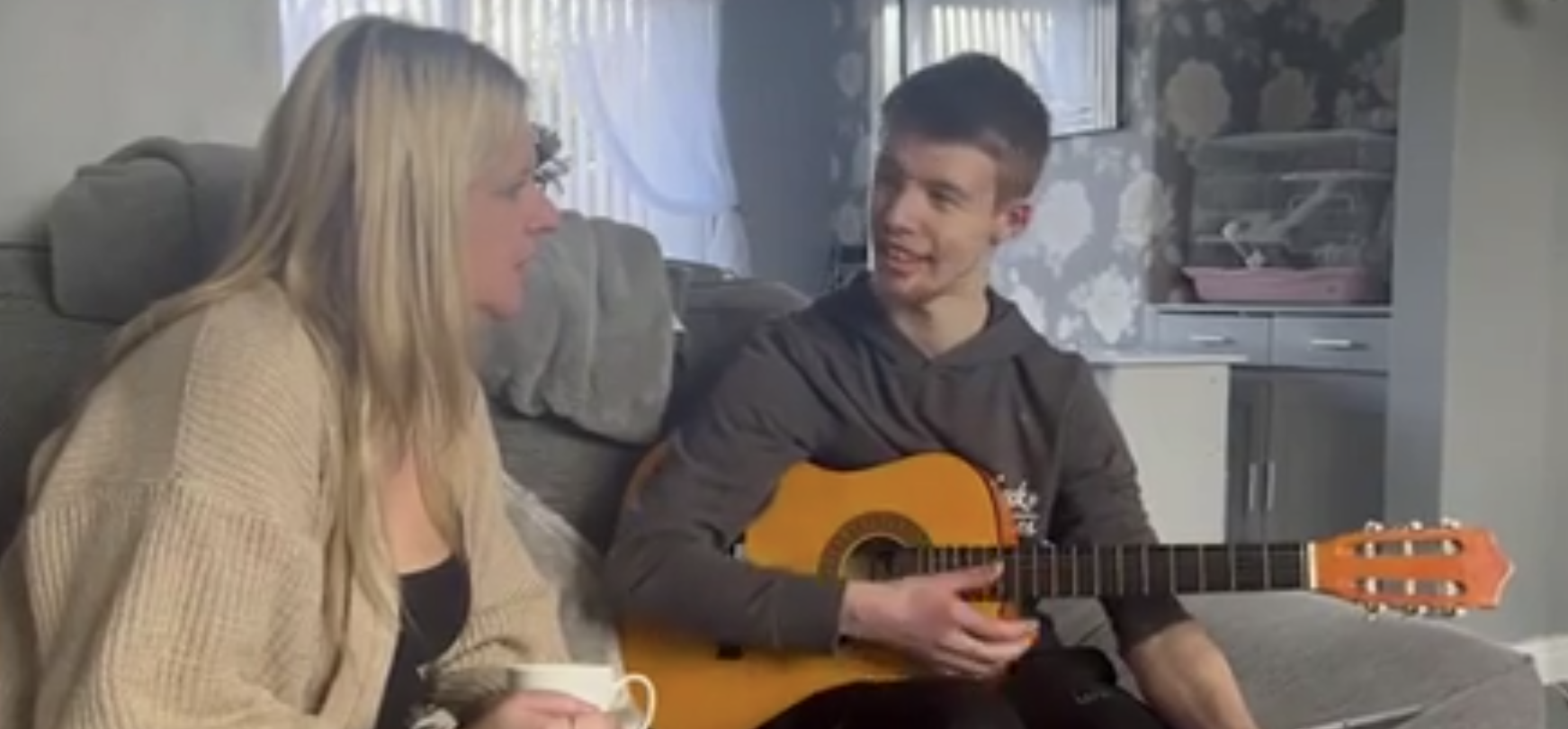 Sara, a blonde adult woman, sits with her back to the camera and her body turned to her teenaged son Josh, who is smiling and holding an acoustic guitar.