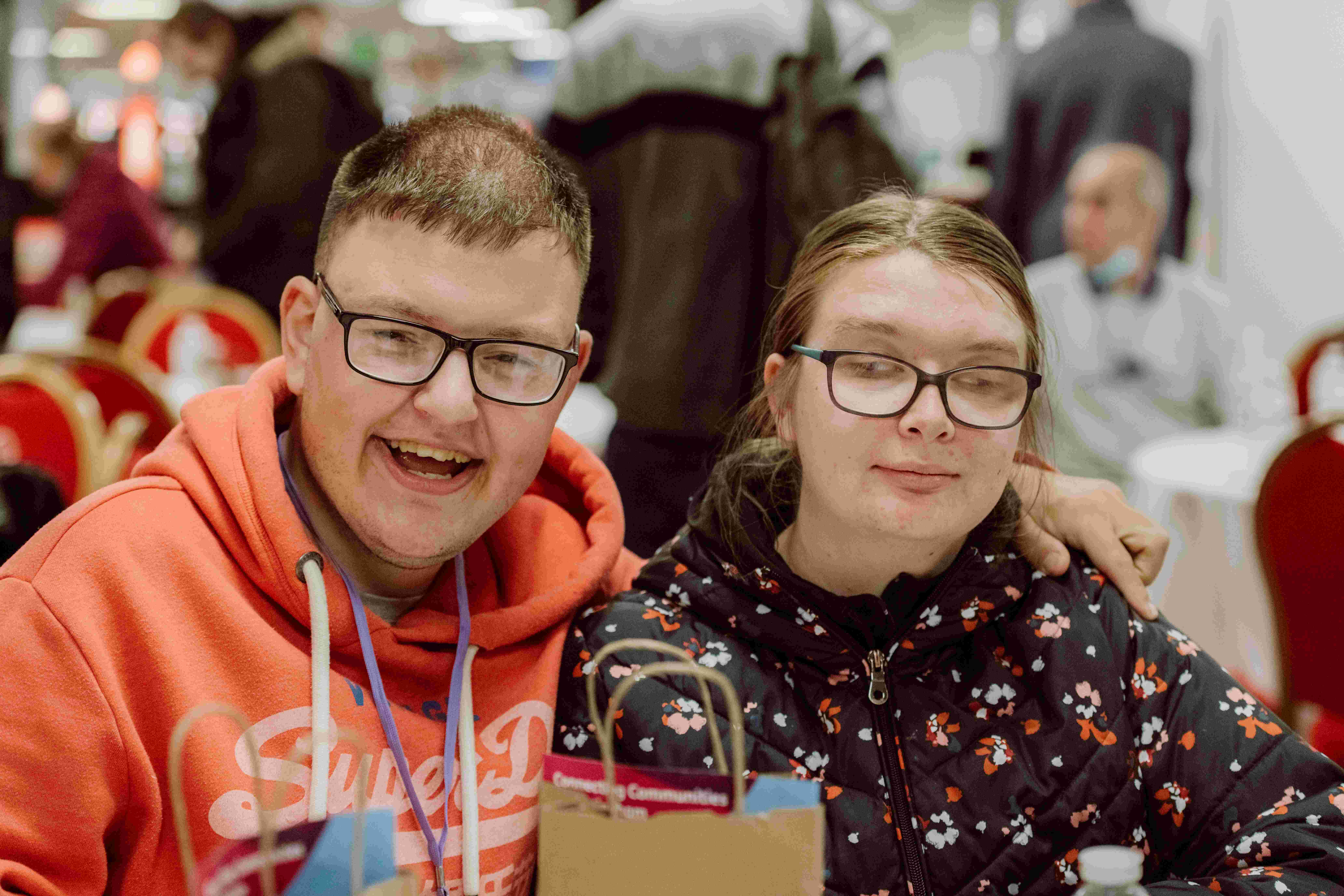 A young man and woman are sat at a table in a community centre smiling at the camera