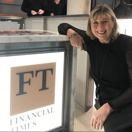 Jacqui has blonde hair and is waring a black suit. She is leaning on sign with the Financial Times logo