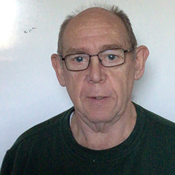 John is standing in front of a white wall indoors. He has rectangular glasses and is wearing a green sweatshirt