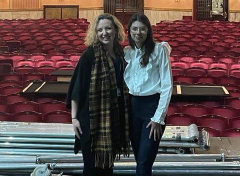 Two women stand close to each other on a stage with rows of theatre seats behind them.