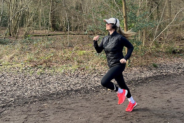 A woman running through woods, with a white baseball cap, black top and trousers, and pink trainers.