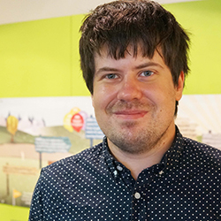 Jack has choppy dark hair and is wearing a blue shirt with a white dotted pattern. He is standing indoors in front of a light green wall