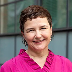 Jackie has short dark hair and is wearing a pink top. She is standing outside with large glass windows behind her.