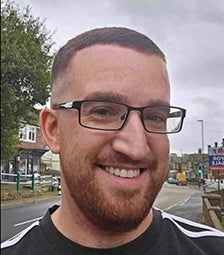 Sam has short dark hair and a beard and is wearing glasses. He is standing outside with a road behind him, and smiling at the camera.