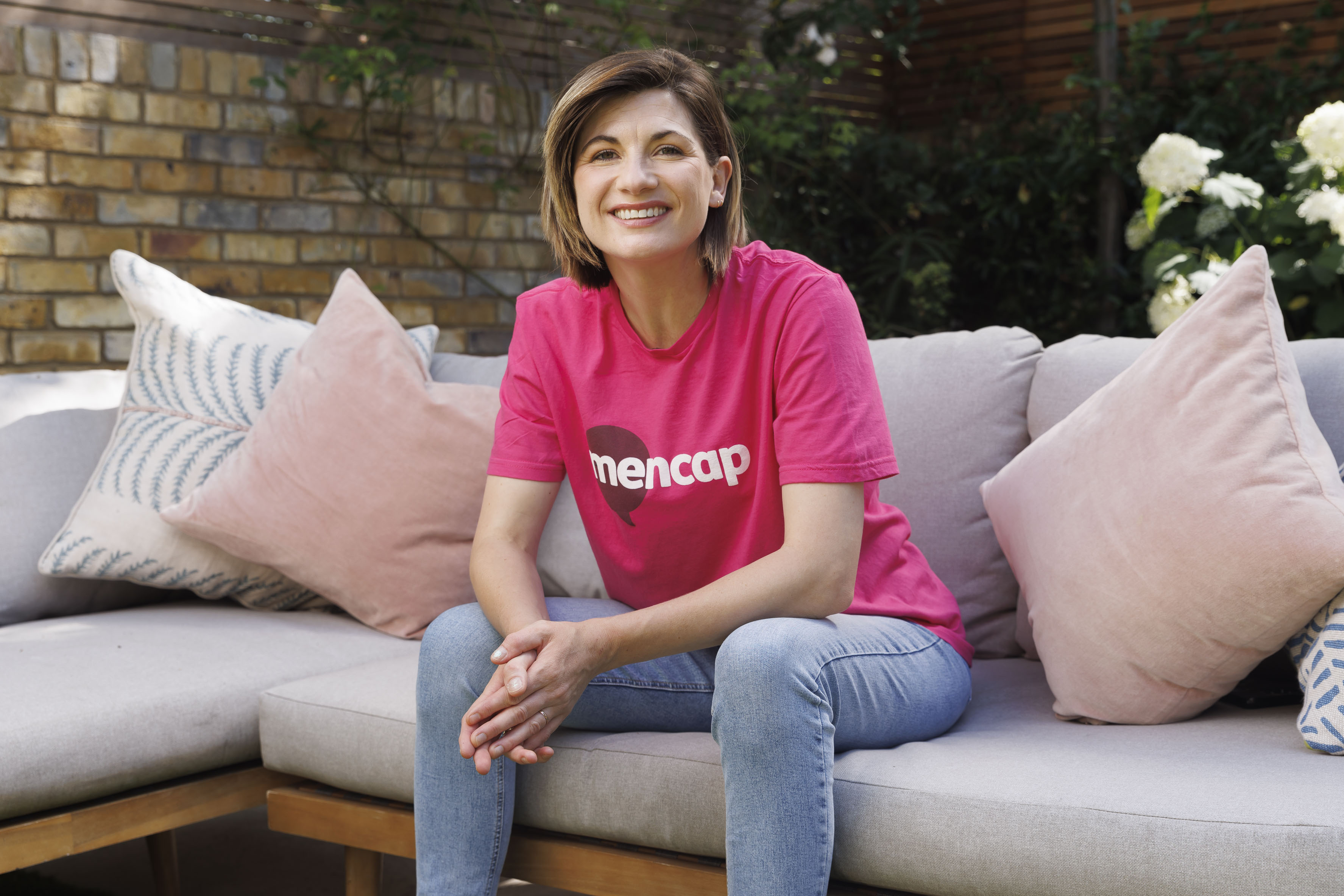 Actress Jodie Whittaker sitting on a sofa wearing a Mencap t-shirt smiling at the camera