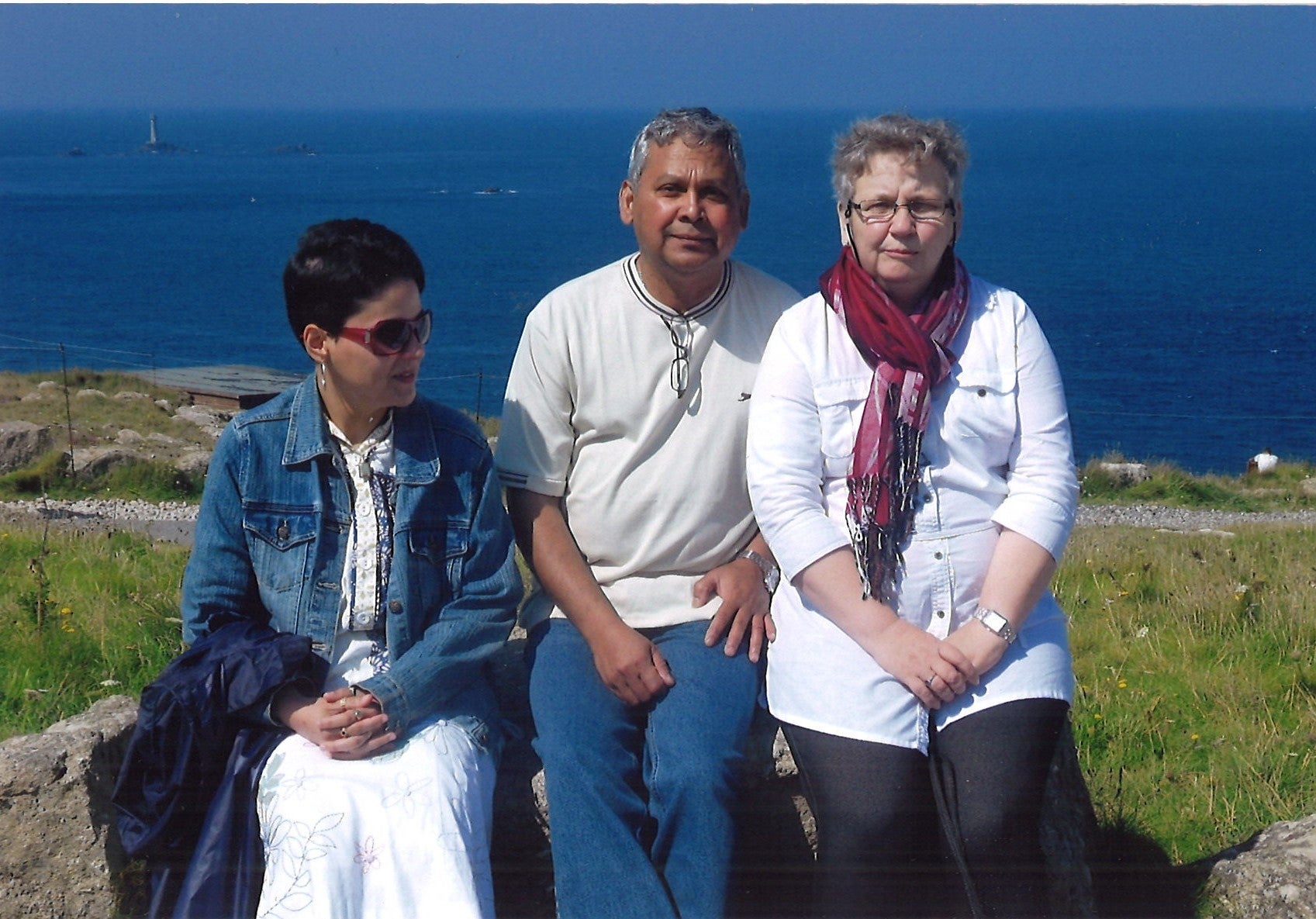 Three people sitting outside on a hill covered with green grass with the sea behind them.