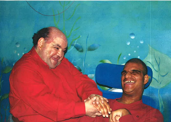 A man in a red shirt stood smiling next to a man in a wheelchair who is also smiling. The wall behind them is decorated with an underwater scene.