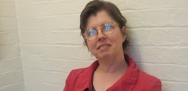 Woman, Amy Clarke, with short dark hair and glasses wearing a red jacket stood in front of white brick wall