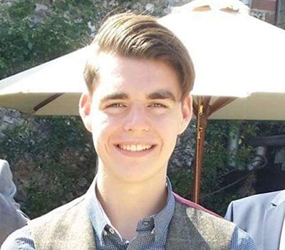 Young man, Oliver, with brown hair stood outside in sunshine smiling, large parasol umbrellas in the background
