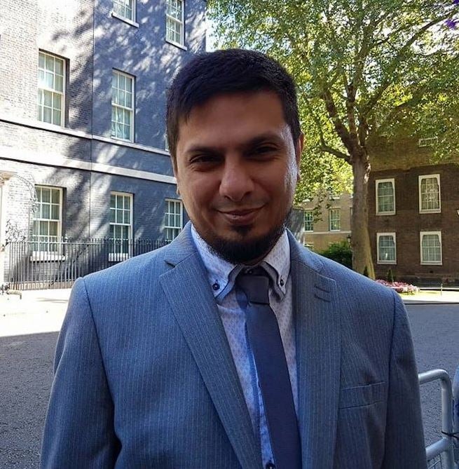 Ismail stands outside 10 Downing Street. He is wearing a grey suit with a blue shirt and tie.