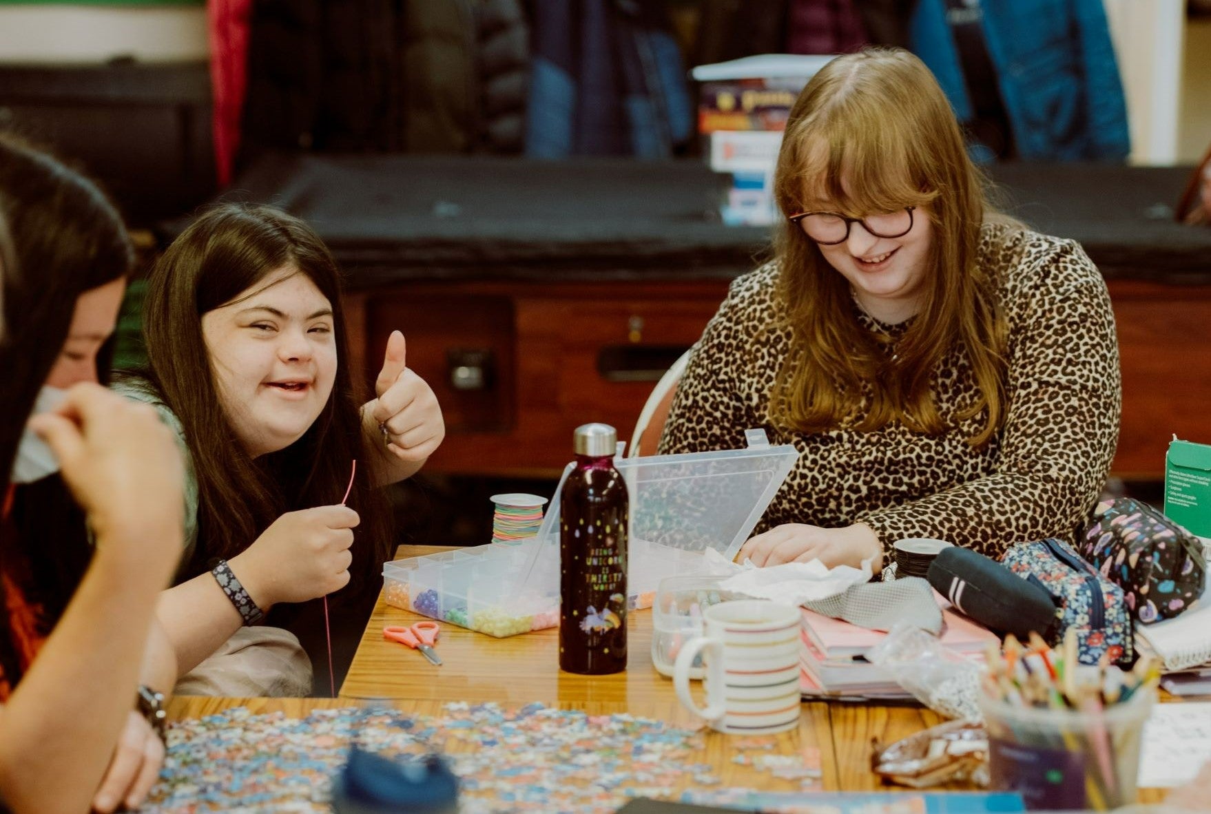 Two women doing artwork at a table. One woman has her thumbs up to the camera and smiling