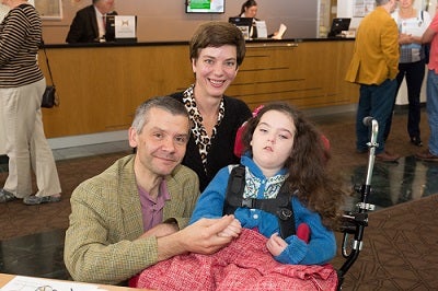 A mum and dad, smiling with their daughter who is in a motorized chair.