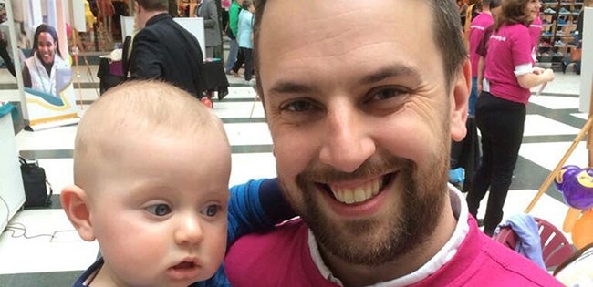 Man, Stephen John, with brown hair and facial hair stood in shopping centre, smiling whilst holding a baby.