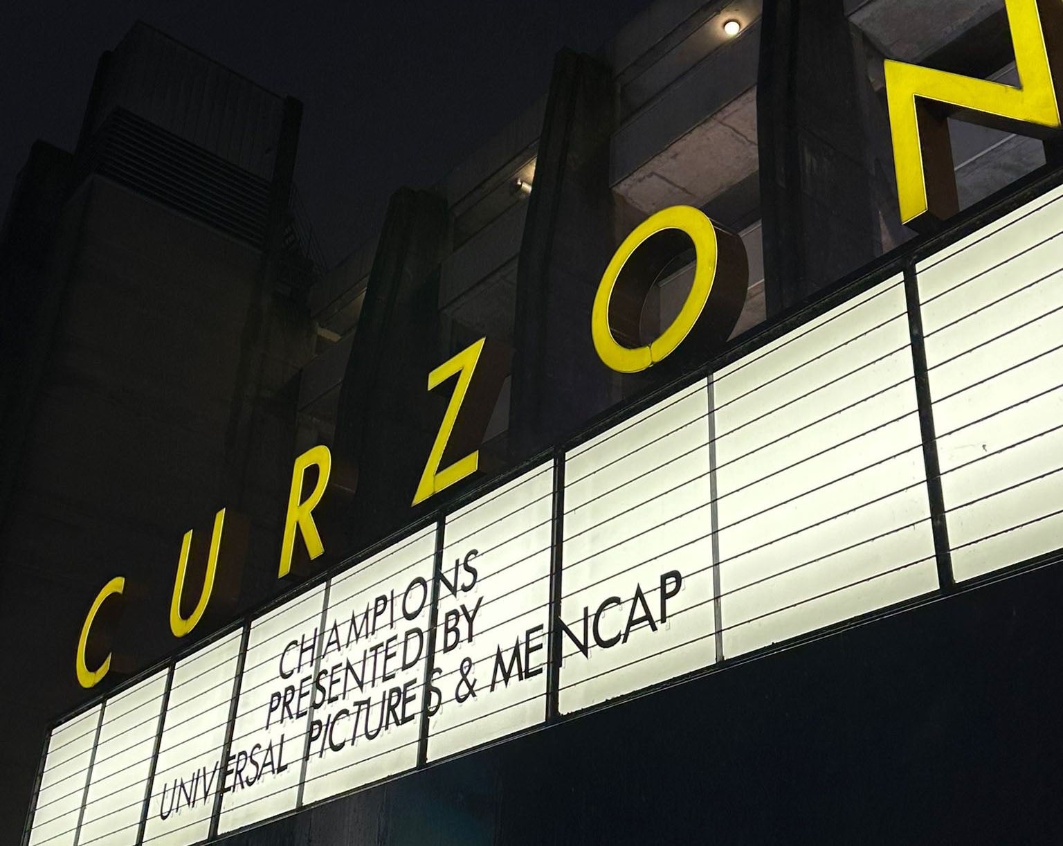 A cinema banner lit up at night, which reads CHAMPIONS PRESENTED BY UNIVERSAL PICTURES & MENCAP