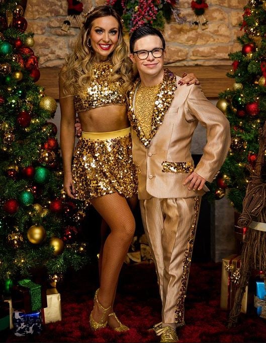Amy Dowden and George Webster stand between two Christmas trees. Amy wears a sparkling gold skirt and crop top. George wears a gold suit with glitter detail on the pockets, lapels and trouser leg.