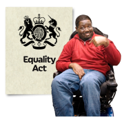 A man in a wheelchair pointing at himself in front of a large poster of the Equality Act