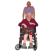 An older lady sitting in a wheelchair wearing slippers and smiling.  A man is standing behind the wheelchair pushing it.