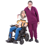 A lady standing who has her arm around a lady next to her in a wheelchair.  They look as if they could be from the same family.