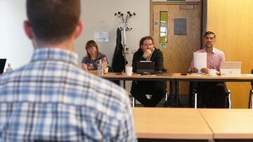 A man at a table with his back to the camera, talking to other people at a meeting who are sitting at an opposite desk.