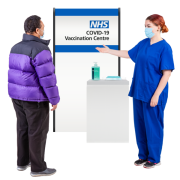 A man wearing a coat is beside a sign which say 'Covid-19 Vaccination Centre'.  A lady wearing a blue uniform and a face mask is looking at him.