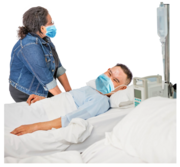A man in a hospital bed wearing a face mask, being visited by a woman also wearing a face mask