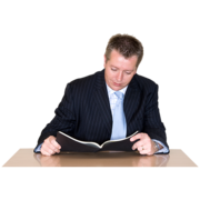 A man in a suit reading a booklet sitting at a desk
