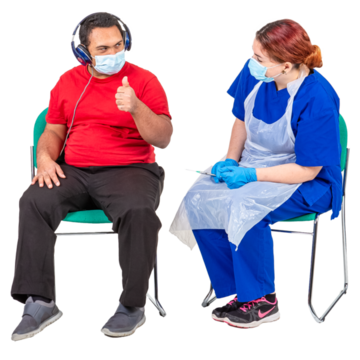 A man sitting on a chair next to a nurse with a needle.  The man is wearing a facemask and headphones and has his thumb up.