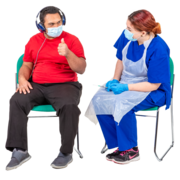 A man sitting on a chair next to a nurse with a needle.  The man is wearing a facemask and headphones and has his thumb up.