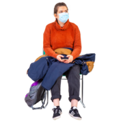 A lady wearing a facemask, sitting on a chair by herself with her phone, coat and bag beside her.