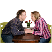 A man and woman face each other holding hands across a table with flowers behind them.