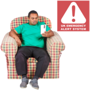 A man sits in an armchair and looks at his phone,  above him is a red box with a triangle warning symbol with text that reads UK Emergency Alert System