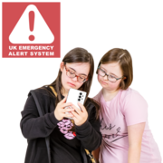 Two women look at a mobile phone. Behind them  is a red box with a triangle warning symbol with text that reads UK Emergency Alert System.