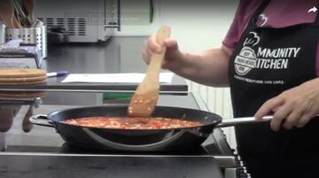 A frying pan of food and a hand holding a wooden spatula.