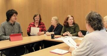 A meeting room where six people are sat around a U shaped meeting table