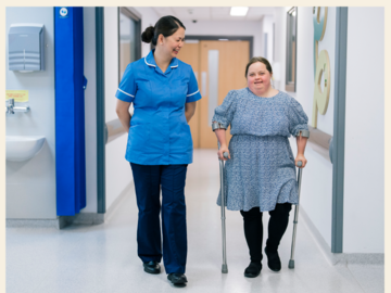 A nurse is walking down a hospital ward with a patient who has Downs Syndrome