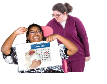 A carer has her arm around a woman in a wheelchair who is holding up a sign which shows the picture of a new born baby and a calendar