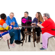 A group of people sat around a table discussing something. There is a man wearing a suit in the middle with a Mencap logo on his jacket.