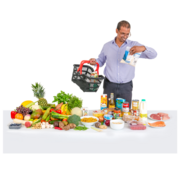 A man carrying a shopping basket is looking a packet of food next to a table full of different foods