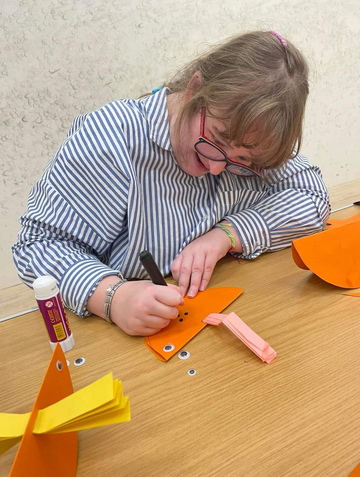 A woman at a table, drawing and sticking things on to a piece of folded paper.