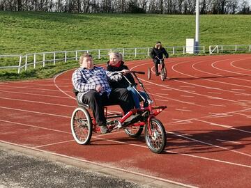 Two people on a two seater bicycle cycle around a running track.