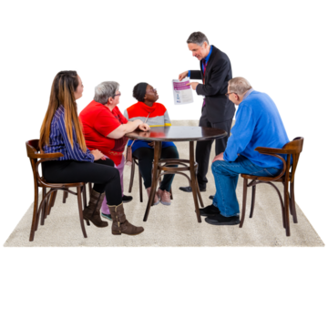 A group of people in a meeting sitting around a desk. One man is standing up showing the others a piece of paper