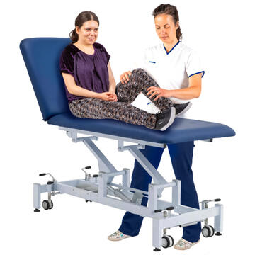 A lady sitting on a therapy couch with a therapist helping her to bend her leg.